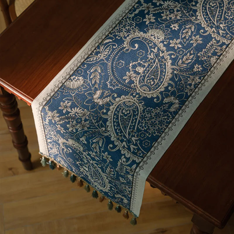 Élégant chemin de table en tissu bleu foncé avec motifs floraux et paisley aux couleurs argentées, orné de glands décoratifs.