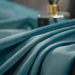 Zoom sur les plis d’un rideau en velours bleu clair illuminé, avec un flacon de parfum en arrière-plan flouté.