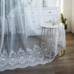 Voilage blanc transparent en 140x200 avec motifs brodés floraux posé dans une chambre lumineuse, tombant délicatement sur un sol en bois foncé.