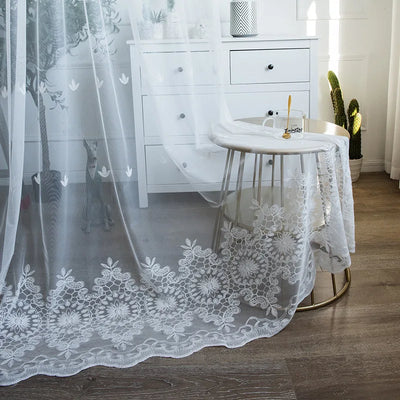Voilage blanc transparent en 140x200 avec motifs brodés floraux posé dans une chambre lumineuse, tombant délicatement sur un sol en bois foncé.