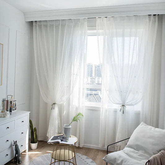 Deux voilages blancs noués avec des embrasses, installés devant une grande fenêtre laissant entrer la lumière naturelle d’un appartement en hauteur.