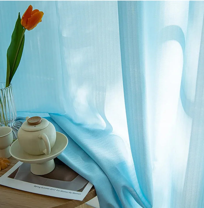 Voilage bleu ciel translucide laissant entrer la lumière naturelle, accompagné d'une tasse de thé et d'une fleur orange en décoration sur une table.