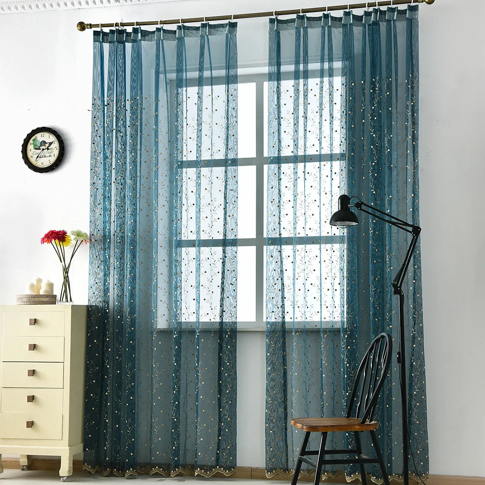 Voilage bleu installé sur une tringle à pattes, laissant passer la lumière, décoré de points brodés discrets.