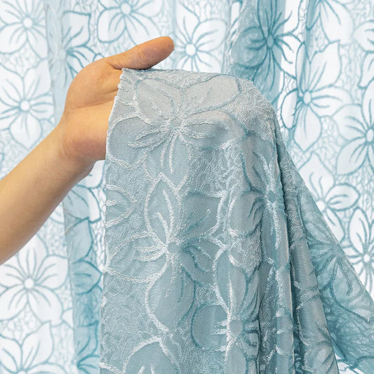 Grosse main tenant un pan de rideau filtrant bleu avec broderie florale, montrant la texture et la finesse du tissu.