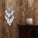 Attrape-rêves en forme de cœur avec plumes blanches suspendu sur un mur en bois rustique, à côté d'un vase décoratif moderne sur une commode. Parfait pour une décoration intérieure apaisante et élégante, ajoutant une touche bohème à votre espace de vie.