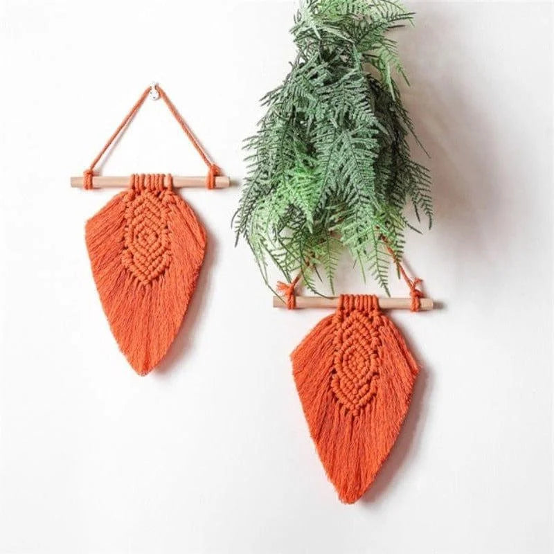 Décoration murale en macramé orange, suspendue avec des bâtons en bois. Idéale pour ajouter une touche bohème à une pièce, elle est accompagnée de verdure pour une ambiance naturelle. Parfaite pour le salon ou la chambre, cette décoration stimule une atmosphère chaleureuse et accueillante.