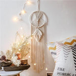 Attrape-rêves bohème décoratif suspendu au mur avec guirlandes lumineuses, accompagné de coussins colorés et d'éléments naturels, créant une ambiance chaleureuse et apaisante pour un intérieur tendance.