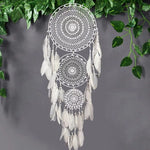 Attrape-rêves décoratif avec motifs détaillés, suspendu devant un mur gris, entouré de feuillage vert. Parfait pour la décoration intérieure bohème ou comme cadeau apaisant pour embellir vos espaces de vie. Cet attrape-rêves ajoute une touche de tranquillité et de style élégant à votre maison.