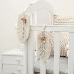 Décoration murale bohème en forme de plume blanche, ornée de perles dans les tons orangés, accrochée à un lit pour bébé. Idéale pour ajouter une touche de douceur et de style dans une chambre d'enfant.