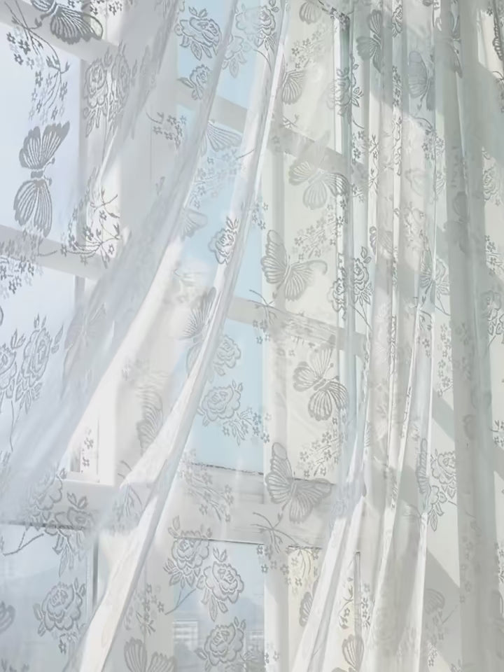 Rideaux voilages blancs imprimés papillons et fleurs installés sur une tringle argentée dans un salon cosy.
