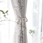 Rideau américain taupe à motifs blancs, attaché avec un cordon torsadé en corde naturelle devant une fenêtre lumineuse.