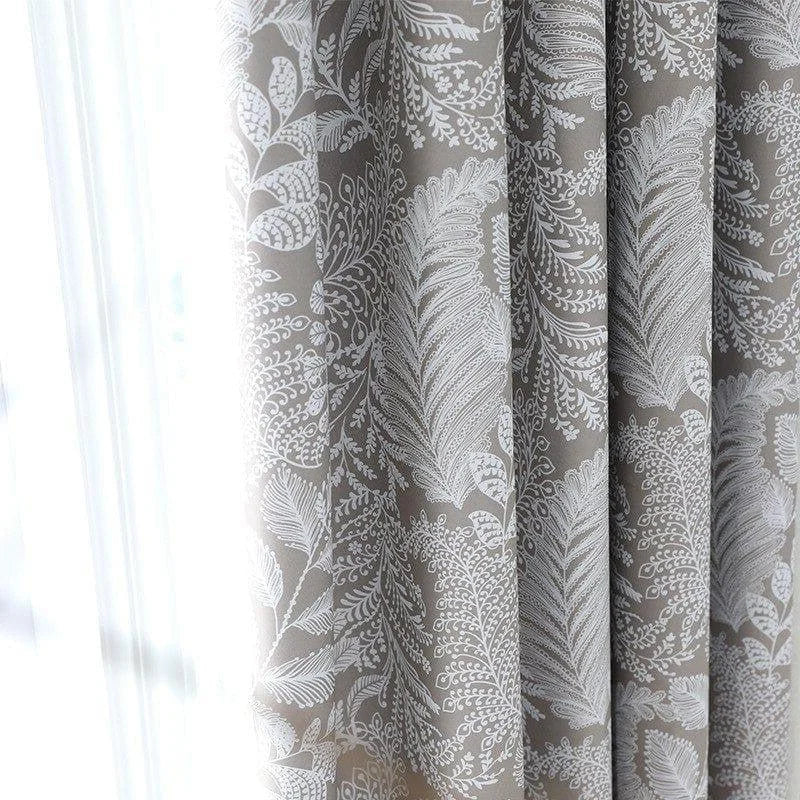 Gros plan sur un rideau américain taupe à motifs blancs végétaux, filtrant la lumière naturelle d'une fenêtre.