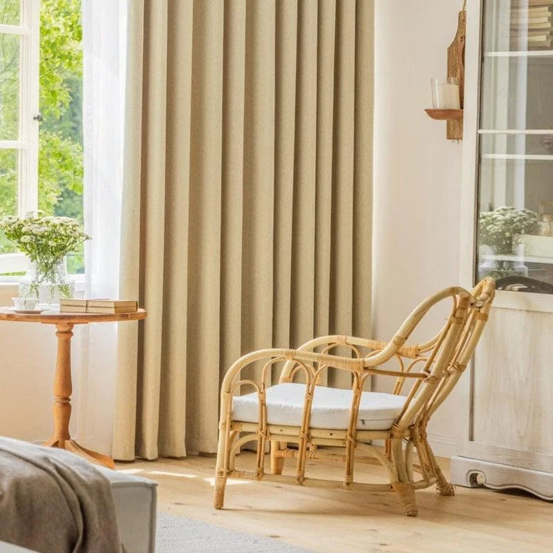 Fauteuil en rotin au design élégant dans un salon lumineux avec des rideaux beiges, une table d'appoint avec des livres et un vase de fleurs, créant une atmosphère chaleureuse et accueillante.