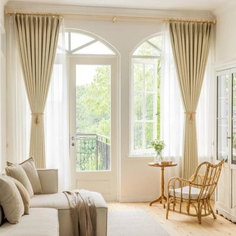 Salon élégant avec rideaux beiges, canapé confortable et chaise en osier, offrant une ambiance lumineuse et chaleureuse. Baie vitrée donnant sur un balcon avec vue sur la nature verdoyante. Aménagement idéal pour une décoration intérieure moderne et sereine.