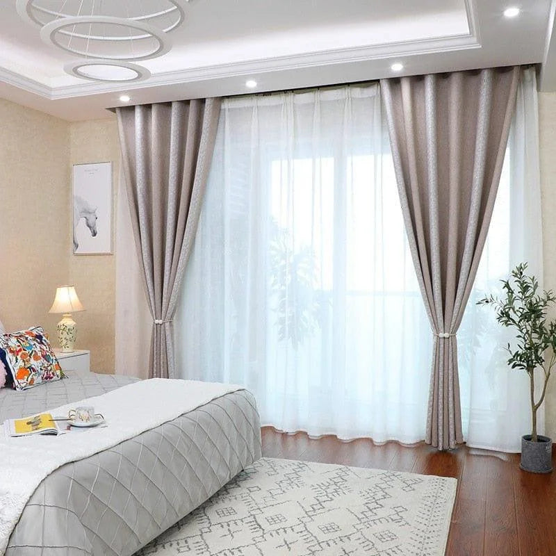 Chambre moderne et élégante avec rideaux opaques gris et voilages blancs, créant une ambiance lumineuse et accueillante. Aménagement raffiné avec un lit confortable, des coussins décoratifs colorés et un tapis géométrique. Parfaite harmonie pour un espace de repos relaxant et stylé.