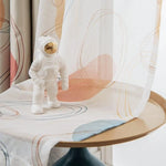 Statue d'astronaute sur une table ronde, entourée de rideaux aux motifs abstraits. Le design moderne de la décoration intérieure ajoute une touche contemporaine à la pièce, avec des couleurs douces et harmonieuses. Idéal pour un salon ou un bureau, apportant une ambiance spatiale et artistique.