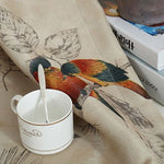 Tasse blanche avec cuillère sur une nappe élégante illustrée de motifs d'oiseaux colorés, posée à côté de livres sur une table de salon. Atmosphère chaleureuse et artistique idéale pour la décoration intérieure.