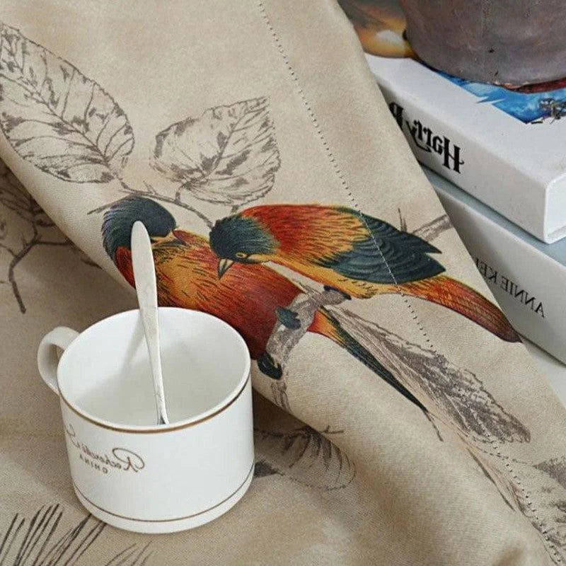 Tasse blanche avec cuillère sur une nappe élégante illustrée de motifs d'oiseaux colorés, posée à côté de livres sur une table de salon. Atmosphère chaleureuse et artistique idéale pour la décoration intérieure.