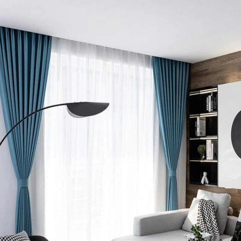 Rideaux élégants en bleu pour salon moderne, avec un design plissé qui ajoute une touche raffinée à la décoration intérieure. Ils encadrent une grande fenêtre avec voilages blancs, permettant une belle lumière naturelle. Parfaits pour un espace de vie lumineux, ces rideaux complètent un mobilier contemporain, associé à une décoration murale minimaliste et des étagères intégrées au style épuré.
