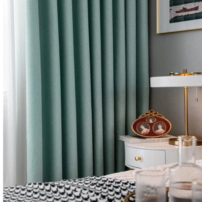 Rideaux opaques vert émeraude ajoutant une touche élégante à la décoration intérieure d'une chambre moderne, harmonieusement associés à une table de chevet blanche, une lampe au design raffiné et une literie avec motifs géométriques noirs et blancs. Ideal pour créer une ambiance cosy et sophistiquée.