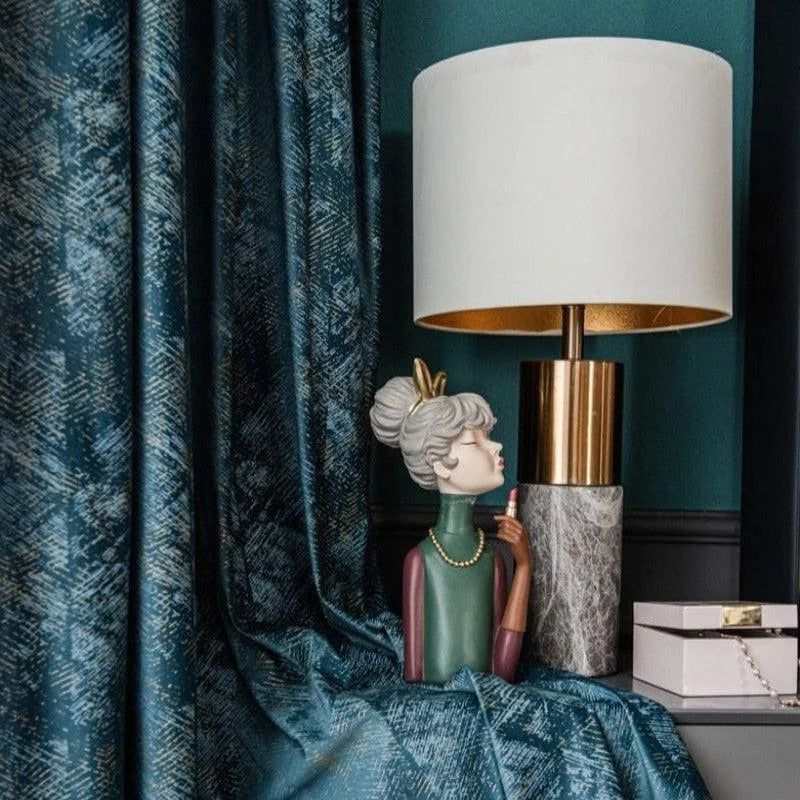 Lampe élégante avec abat-jour blanc et base dorée, posée sur une table d'appoint. À ses côtés, une statuette décorative colorée représente une femme stylisée avec un collier. Rideaux bleu profond avec motifs texturés en arrière-plan, ajoutant une touche de sophistication à l'ensemble du décor. Cette composition raffinée est parfaite pour une décoration intérieure moderne et chic.