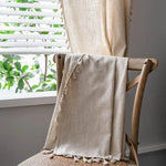 Rideau beige avec franges décoratives posé sur une chaise en bois près d'une fenêtre à persiennes blanches, créant une atmosphère lumineuse et naturelle dans la pièce. Idéal pour ajouter une touche d'élégance à votre intérieur tout en préservant l'intimité. Accessoire parfait pour un salon ou une chambre, apportant chaleur et confort.