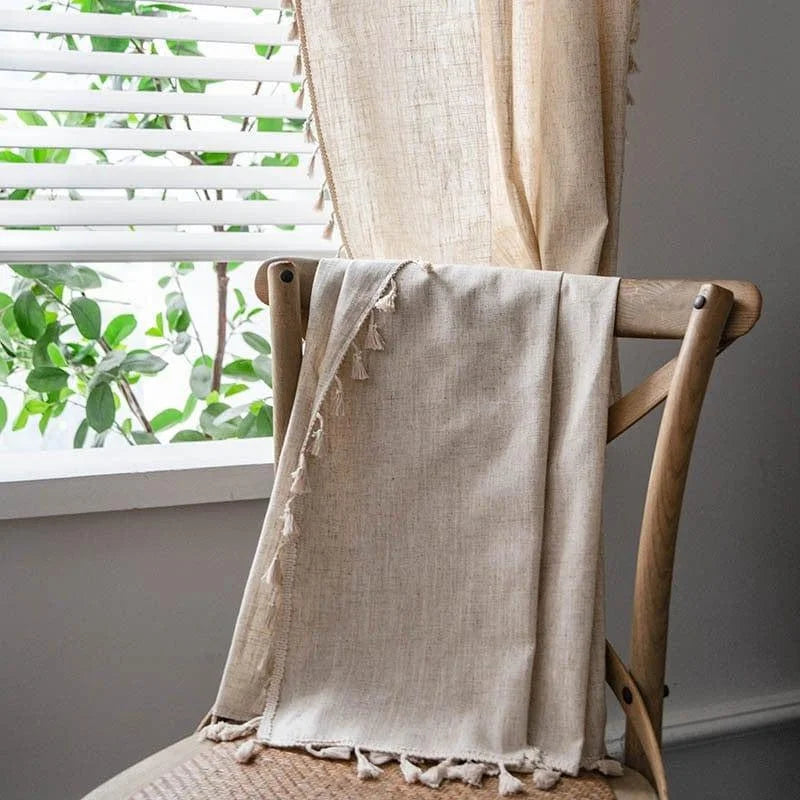 Rideau beige avec franges décoratives posé sur une chaise en bois près d'une fenêtre à persiennes blanches, créant une atmosphère lumineuse et naturelle dans la pièce. Idéal pour ajouter une touche d'élégance à votre intérieur tout en préservant l'intimité. Accessoire parfait pour un salon ou une chambre, apportant chaleur et confort.