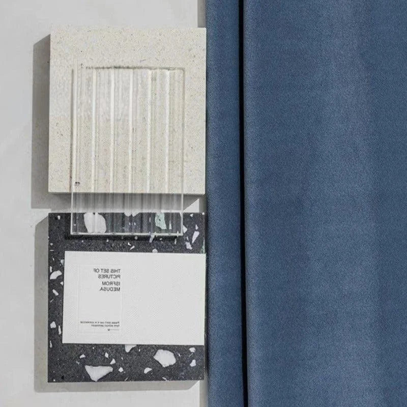 Décor moderne avec échantillons de différentes textures et couleurs. Bloc blanc nuancé, élément transparent à rayures verticales, et panneau noir moucheté de blanc. Rideau bleu à droite, ajoutant une touche élégante à l'ensemble. Idéal pour l'inspiration design intérieur et choix de matériaux.