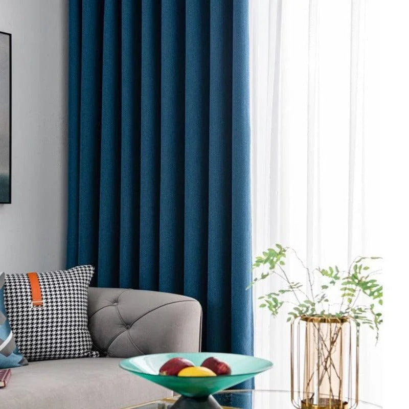 Rideau bleu élégant dans un salon moderne, accentué par un canapé gris avec des coussins à motifs géométriques, une table basse en verre avec un plateau décoratif et une plante verte en arrière-plan. Idéal pour ajouter une touche contemporaine et sophistiquée à votre décoration intérieure.