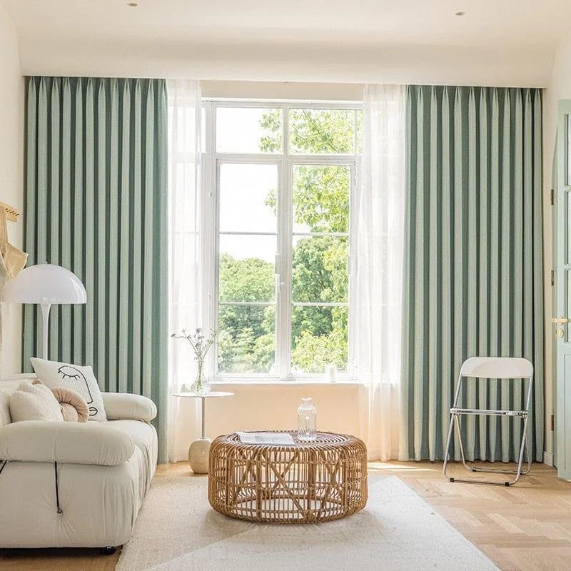 Salon lumineux avec de grands rideaux verts, canapé confortable, table basse tressée et décoration minimaliste. Fenêtre ouverte sur un jardin verdoyant en arrière-plan, créant une ambiance naturelle et chaleureuse. Idéal pour un intérieur moderne et serein.