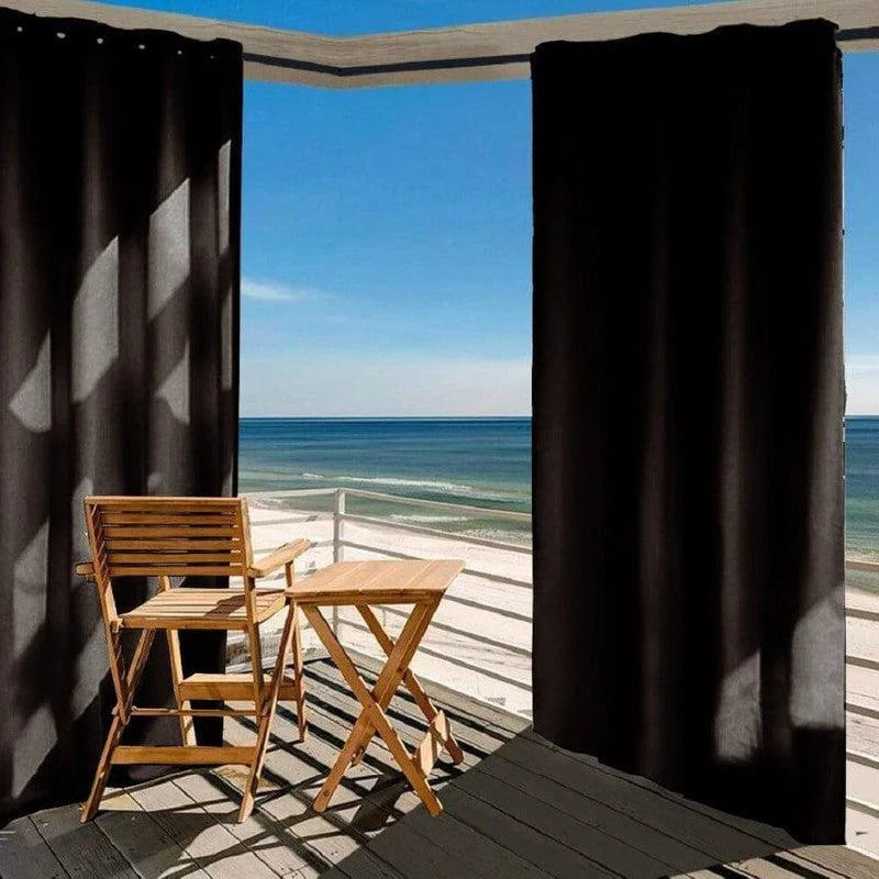 Voilage occultant noir pour terrasse avec vue sur la mer, créant un espace extérieur ombragé et intime. Ensemble de mobilier en bois léger idéal pour les moments de détente en plein air. Parfait pour créer une ambiance élégante et pratique sur un balcon ou une véranda.