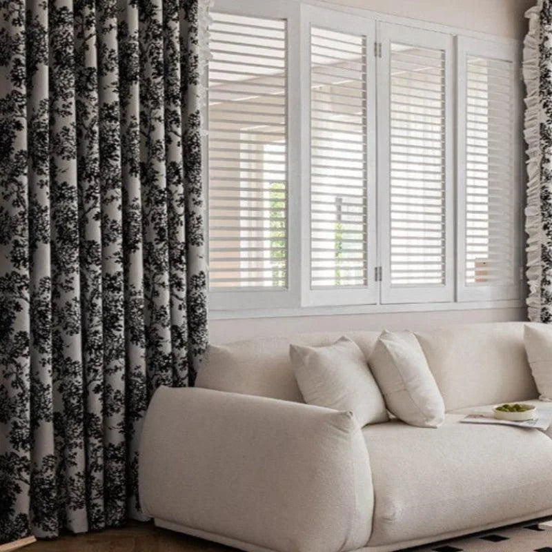 Canapé moderne beige avec coussins, devant rideaux élégants à motif floral noir et blanc, offrant un style chic et contemporain à un salon lumineux. Fenêtres à lamelles ajoutent une touche de sophistication et optimisent la lumière naturelle. Idéal pour une décoration intérieure accueillante et raffinée.