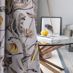 Rideau à motif floral tropical avec des citrons en décoration, suspendu dans une salle à manger contemporaine. Table en verre avec base en bois moderne et un citron posé sur des magazines. Ambiance lumineuse et élégante idéale pour un intérieur raffiné.