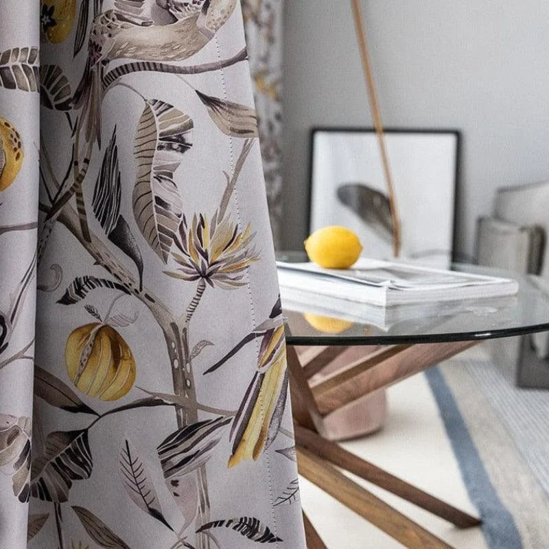 Rideau à motif floral tropical avec des citrons en décoration, suspendu dans une salle à manger contemporaine. Table en verre avec base en bois moderne et un citron posé sur des magazines. Ambiance lumineuse et élégante idéale pour un intérieur raffiné.