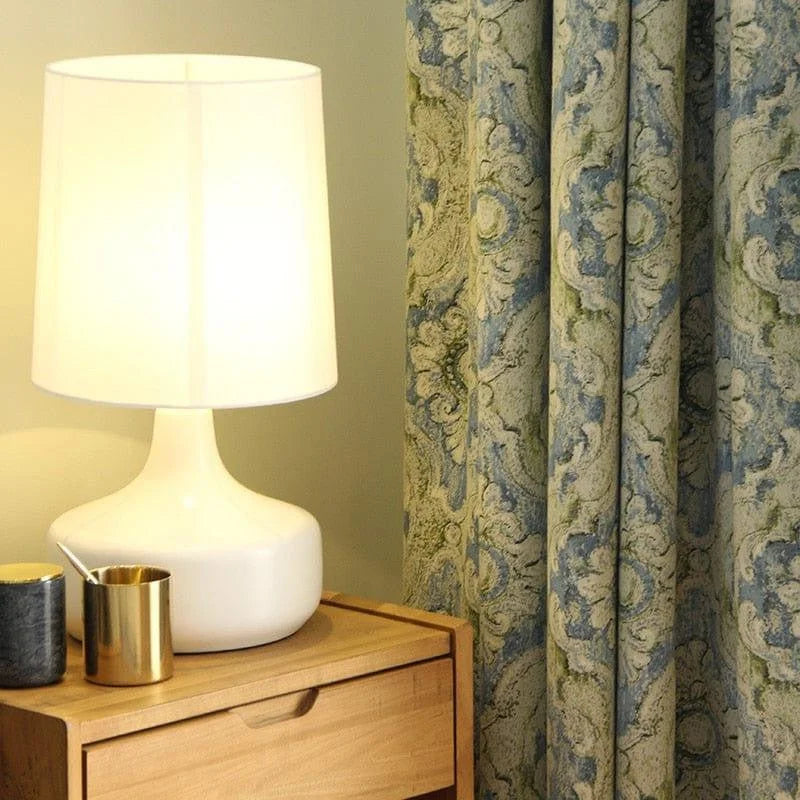 Lampe de table moderne avec abat-jour blanc posée sur une table en bois clair, accompagnée d'un rideau à motifs élégants en arrière-plan. Idéale pour ajouter une touche de sophistication à votre salon ou chambre à coucher. Accessoires de décoration discrets mais chic visibles sur la table.