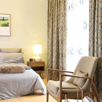 Chambre élégante avec lit douillet et fauteuil confortable près de la fenêtre, éclairée par une lampe de chevet moderne. Rideaux à motifs floraux apportant une touche chic et chaleureuse à la décoration. Espace harmonieux et accueillant idéal pour la détente et le repos.