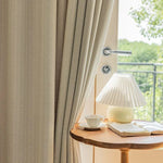 Rideau beige élégant suspendu à une fenêtre lumineuse, offrant une vue sur la nature environnante, accompagné d'une lampe design posée sur une table en bois, créant une ambiance chaleureuse et accueillante. Découvrez comment transformer votre intérieur avec style et luminosité.