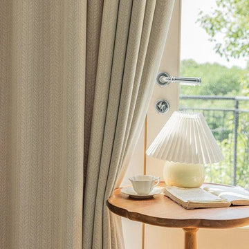 Rideau beige élégant suspendu à une fenêtre lumineuse, offrant une vue sur la nature environnante, accompagné d'une lampe design posée sur une table en bois, créant une ambiance chaleureuse et accueillante. Découvrez comment transformer votre intérieur avec style et luminosité.