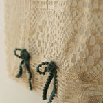 Image de gros plan d'un textile beige ajouré avec des motifs complexes, décoré de petits nœuds verts pour une touche élégante et raffinée, idéale pour les projets de design intérieur ou la confection de vêtements sophistiqués.