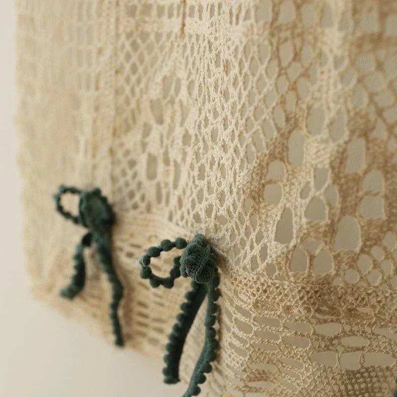 Image de gros plan d'un textile beige ajouré avec des motifs complexes, décoré de petits nœuds verts pour une touche élégante et raffinée, idéale pour les projets de design intérieur ou la confection de vêtements sophistiqués.