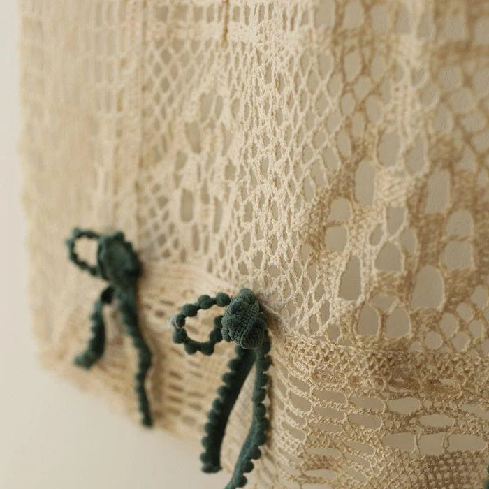 Image de gros plan d'un textile beige ajouré avec des motifs complexes, décoré de petits nœuds verts pour une touche élégante et raffinée, idéale pour les projets de design intérieur ou la confection de vêtements sophistiqués.