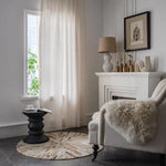 Salon élégant avec fauteuil blanc confortable et table d'appoint noire, décor moderne comprenant une lampe et des objets décoratifs dorés sur une cheminée blanche, atmosphère lumineuse avec rideaux transparents et lumière naturelle.
