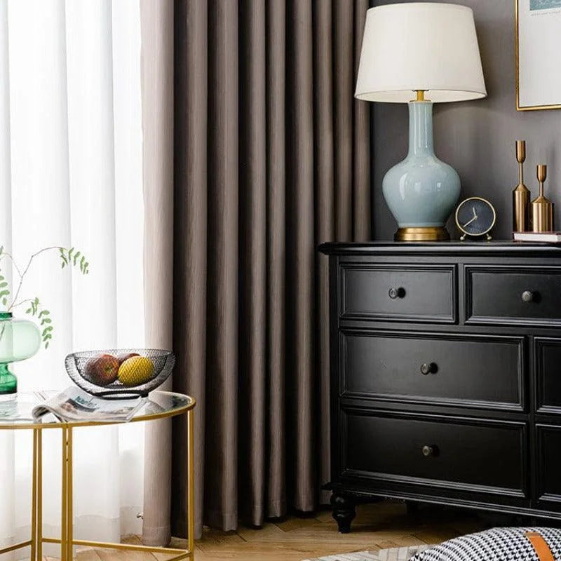 Table d'appoint dorée avec plateau en verre, accompagnée d'une lampe de table moderne sur une commode noire. Rideaux élégants en arrière-plan et accessoires décoratifs ajoutant une touche de sophistication à la pièce. Ambiance chaleureuse et chic pour un intérieur contemporain.