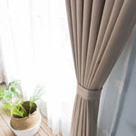 Rideau élégant beige fermé par une attache, offrant une ambiance moderne et apaisante dans un salon lumineux. Décoration intérieure avec un cache-pot tressé et une plante verte sur un sol en bois. Idéal pour filtrer la lumière tout en ajoutant une touche de style à votre espace de vie.