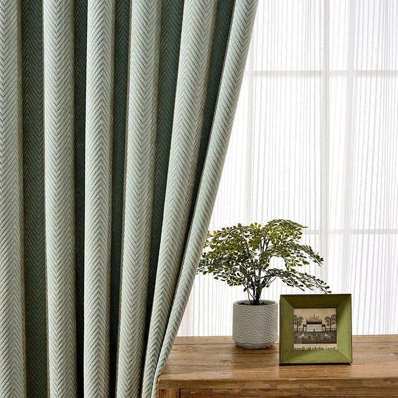 Rideau vert au motif chevron, apportant une touche élégante et moderne à votre intérieur. Idéal pour filtrer la lumière tout en ajoutant du style à votre salon ou chambre. Complément parfait d'une décoration contemporaine, avec un joli contraste sur une table en bois ornée d'une plante en pot.