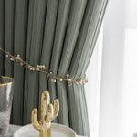Rideau élégant vert clair, attaché par un cordon décoratif orné de perles blanches, ajoutant une touche sophistiquée et moderne à la décoration intérieure. Accessoires dorés en avant-plan, mettant en valeur le design luxueux pour un salon ou une chambre à coucher. Style contemporain et raffiné adapté à tous les environnements de maison.