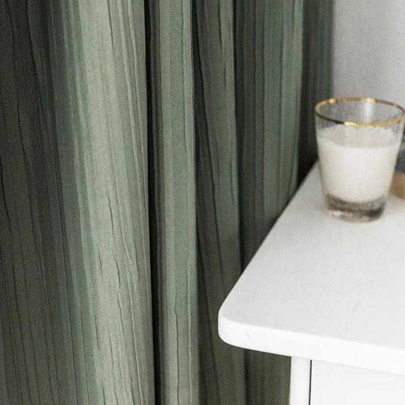 Rideaux verts élégants tombant harmonieusement à côté d'une table blanche moderne avec un verre vide, apportant une touche de sophistication à la décoration intérieure.