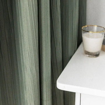 Rideaux verts élégants tombant harmonieusement à côté d'une table blanche moderne avec un verre vide, apportant une touche de sophistication à la décoration intérieure.