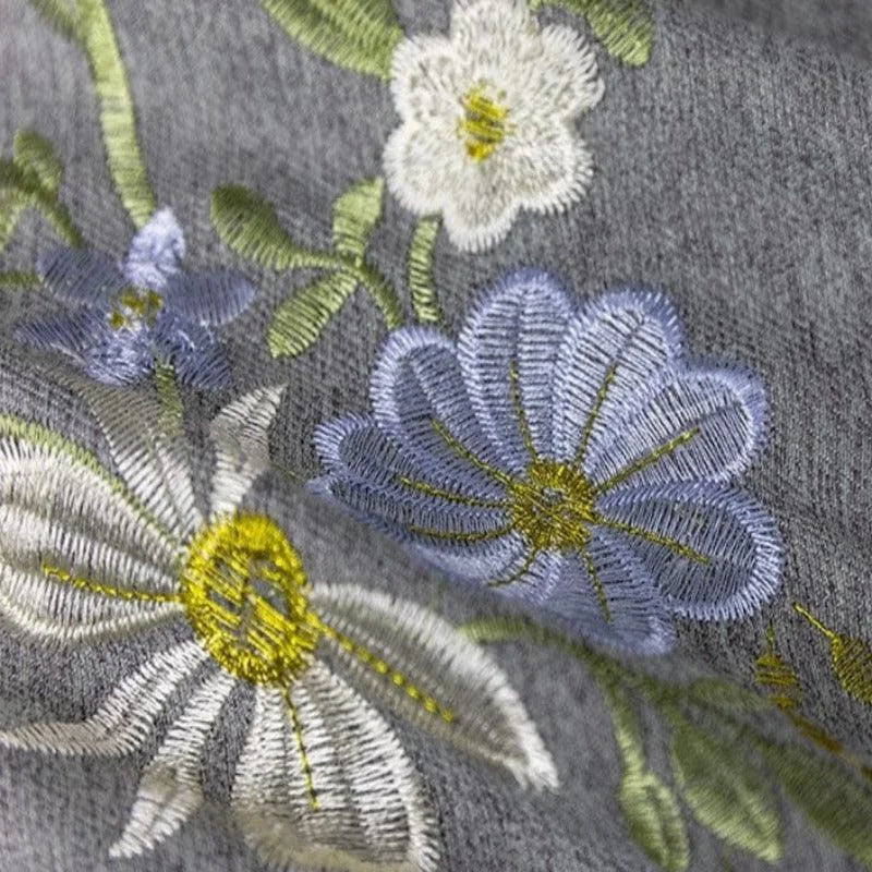 Broderie florale délicate sur tissu gris, mettant en valeur des motifs de fleurs blanches et bleues avec des détails jaunes et verts, idéal pour ajouter une touche élégante à vos créations textiles.
