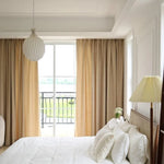 Chambre lumineuse et élégante avec lit double, rideaux beiges et lampe de chevet. Fenêtre donnant sur un balcon avec vue sur la nature. Conception harmonieuse pour une ambiance relaxante et moderne.